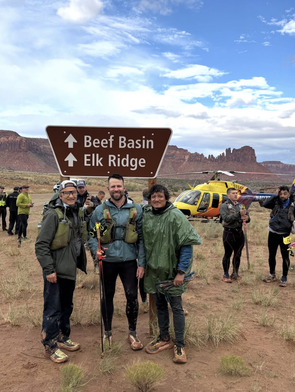 Three runners in muddy shoes at the Beef Basin and Elk Ridge sign, with a yellow helicopter and other runners behind them